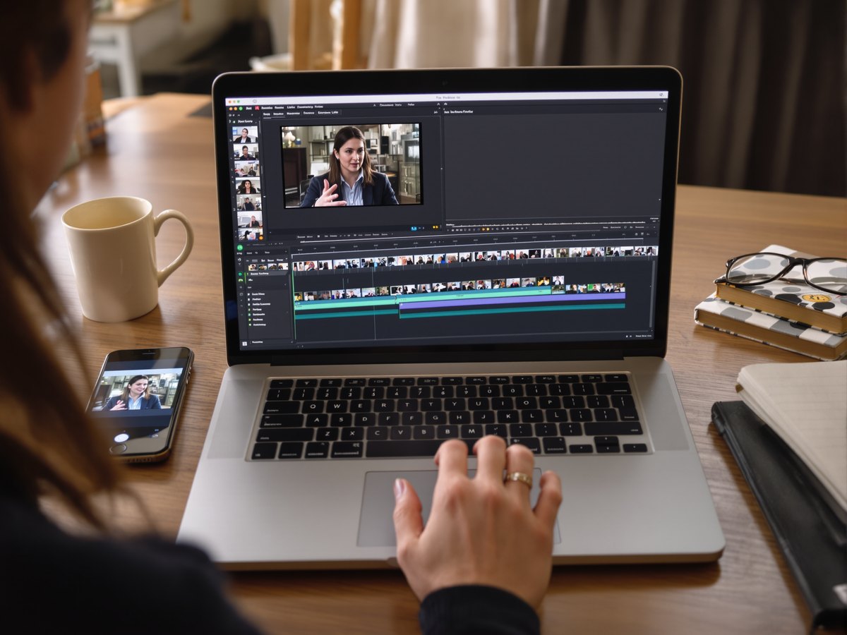 A laptop on a desk showing a video editing timeline, with the same clip playing on a phone beside it